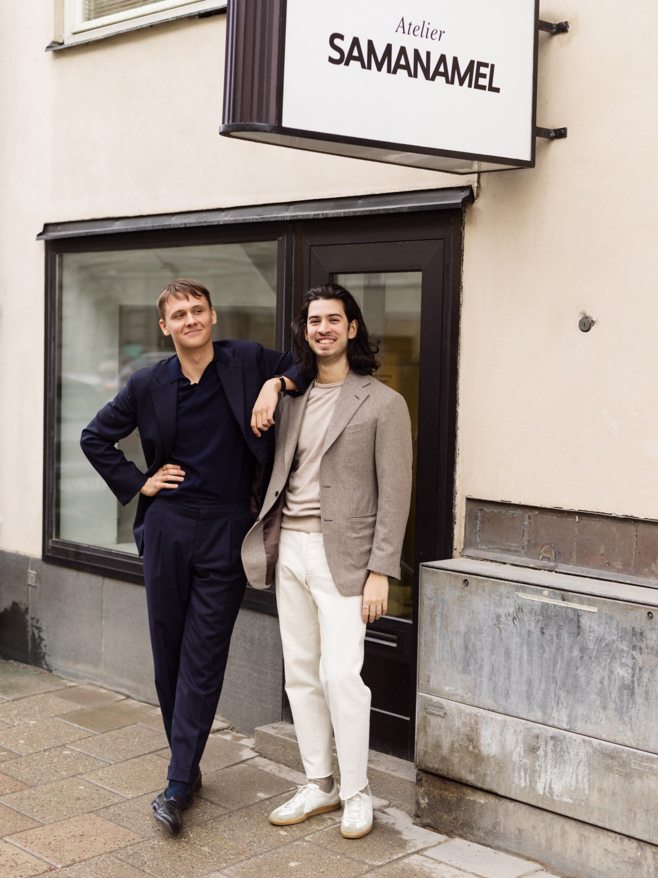 Two men stand outside Atelier Samanamel, one wearing a dark navy suit, the other in a beige blazer with cream trousers.