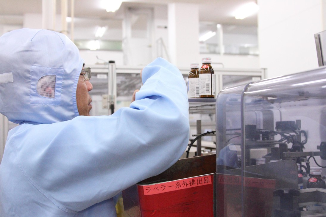 Quality control worker in sterile white protective clothing inspecting a vial at Taisho Pharmaceutical manufacturing facility.