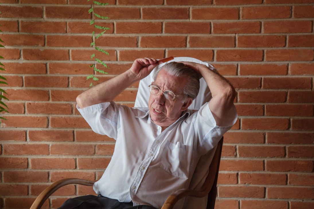 An elderly man in a white shirt and glasses stretches against a brick wall with ivy