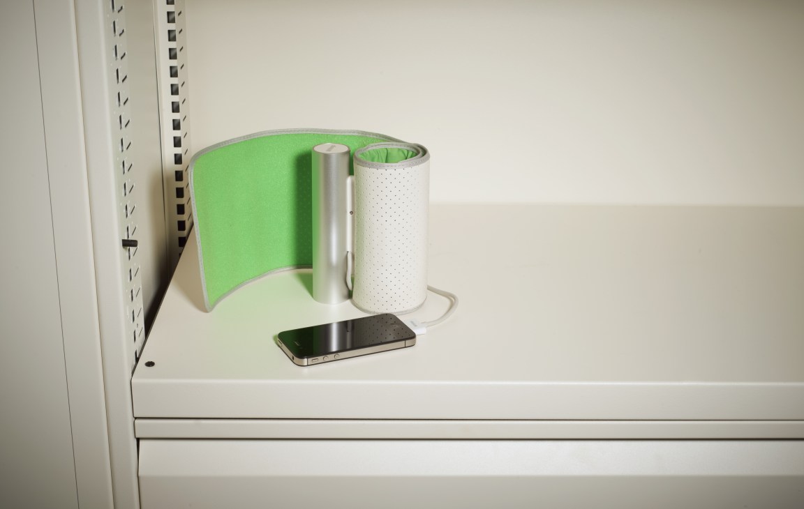 Withings blood pressure monitor armband in green with silver casing on white shelf next to smartphone.