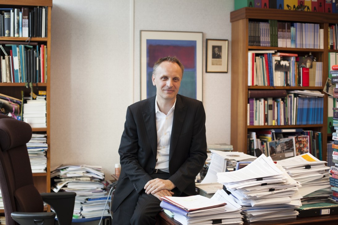 Richard Horton, editor-in-chief of The Lancet, sits at his desk surrounded by stacks of papers and books with bookshelves behind him.