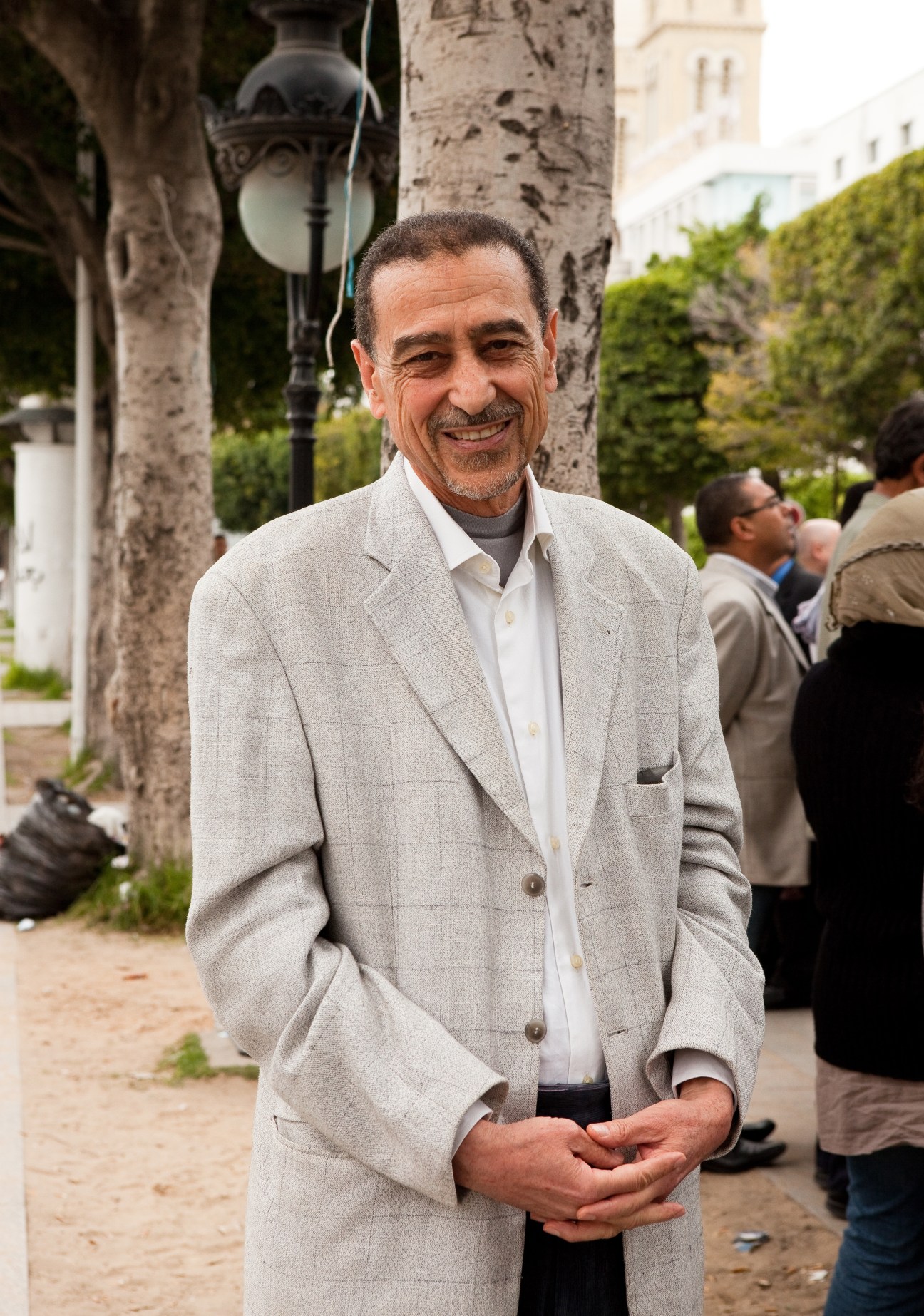Man in light beige blazer and white shirt standing outdoors on a tree-lined street in Tunisia.