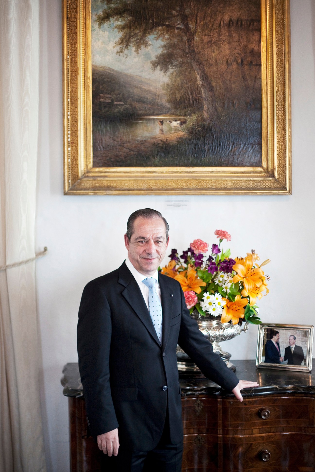 Prime Minister Lawrence Gonzi in a dark suit standing in an elegant interior with framed artwork and flowers.