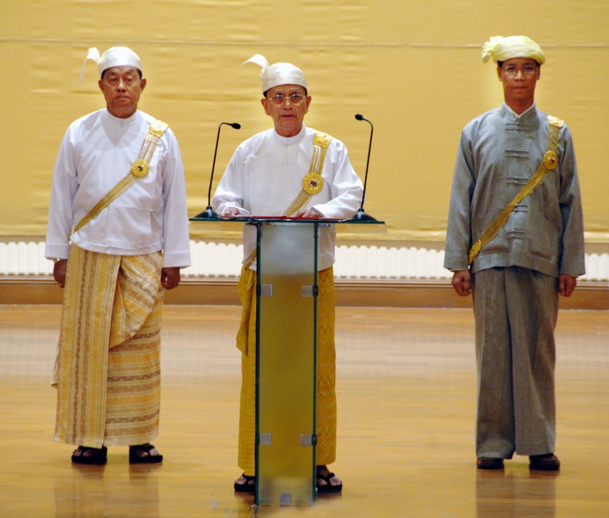 Thein Sein stands at a podium in traditional Burmese dress, flanked by two uniformed officials in a golden-walled parliament hall.