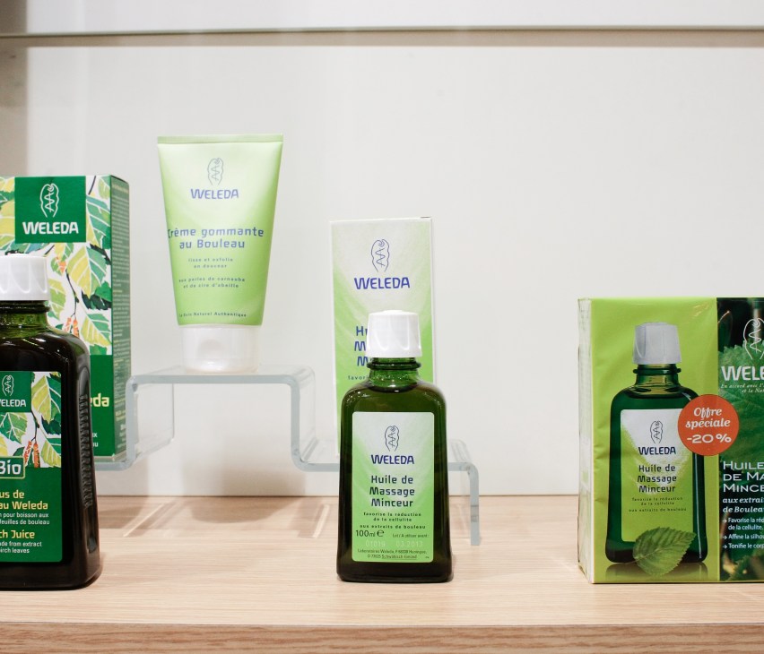 Weleda skincare and wellness products displayed on a wooden shelf, including massage oil, creams, and packaged items.
