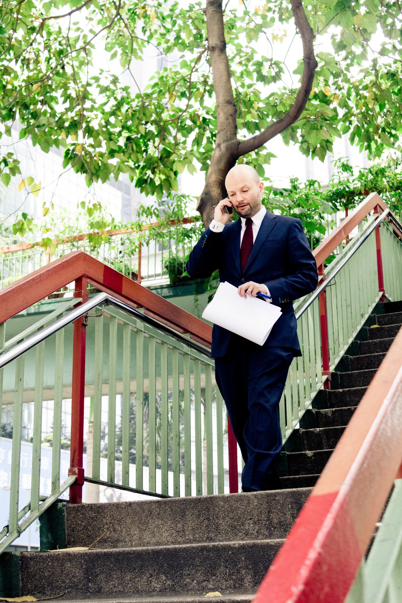 Man in dark suit walking down stairs while talking on phone, holding a folder, beneath tree foliage.