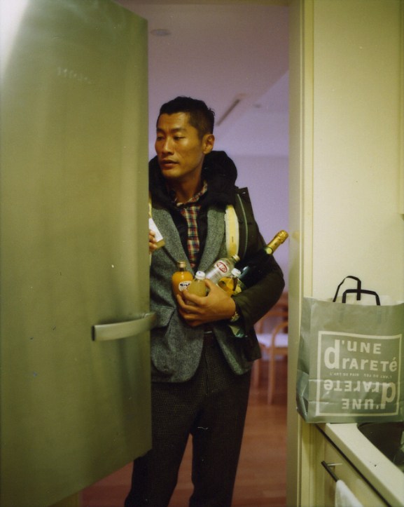 Man wearing Visvim blouson jacket, Tokito jacket, and Bottega Veneta trousers holds a yellow bag indoors.