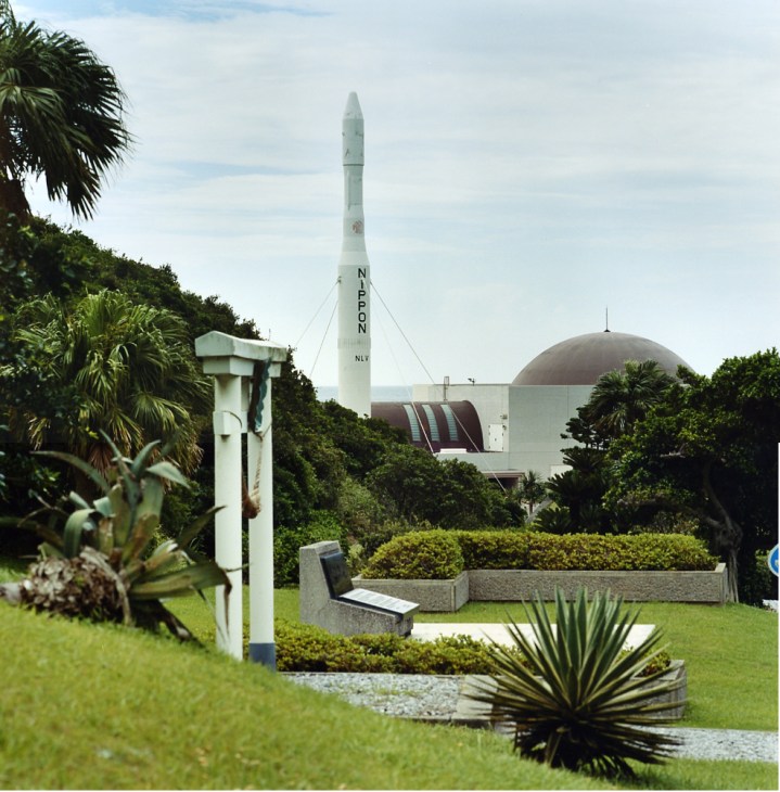 A white rocket model stands tall beside a white domed space museum building surrounded by tropical vegetation.