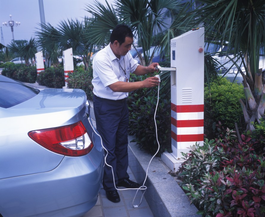 Man in white shirt charging a blue electric car at a white and red charging station surrounded by tropical plants.