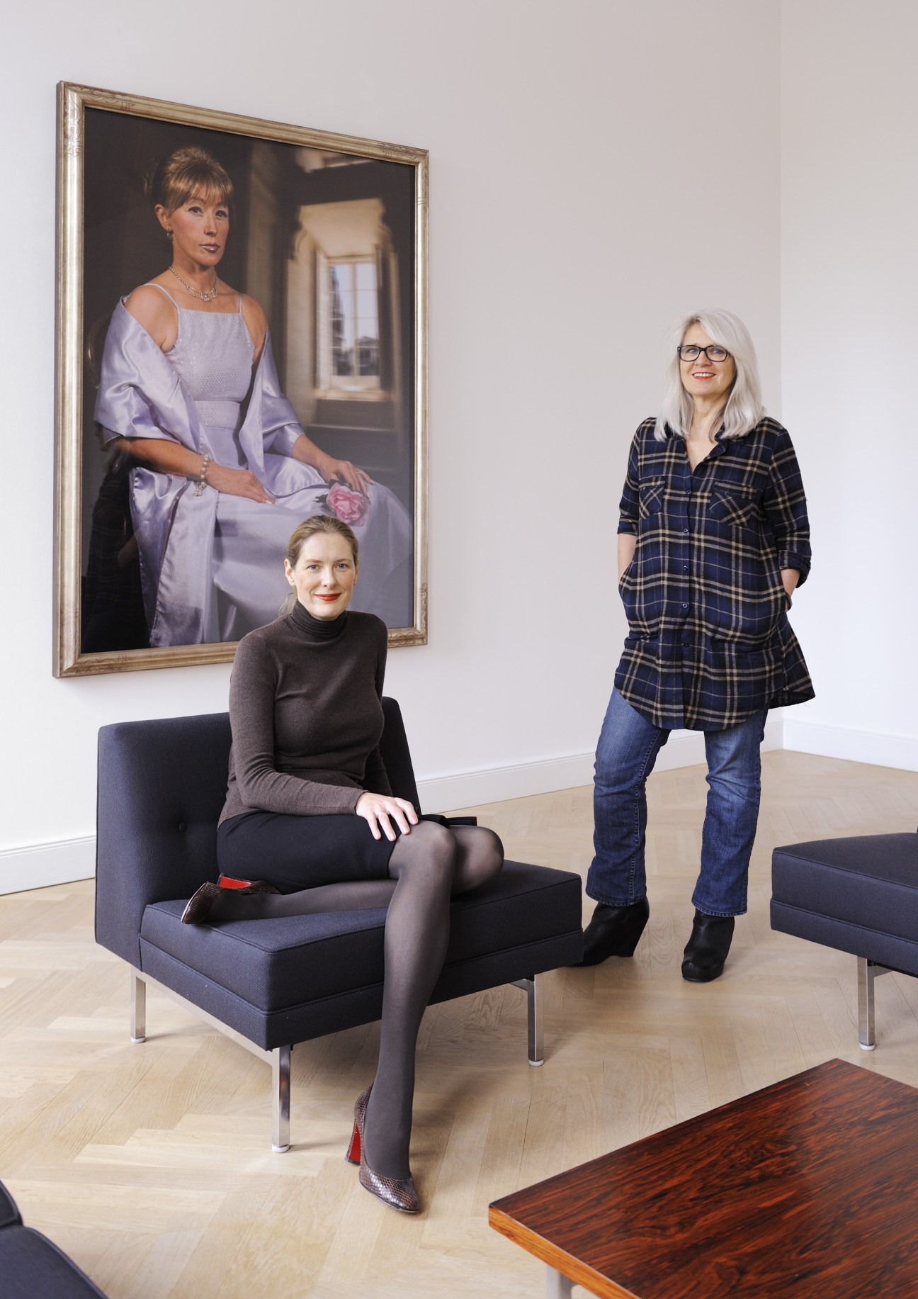 Two women in a modern gallery space with a framed portrait painting on the wall behind them.