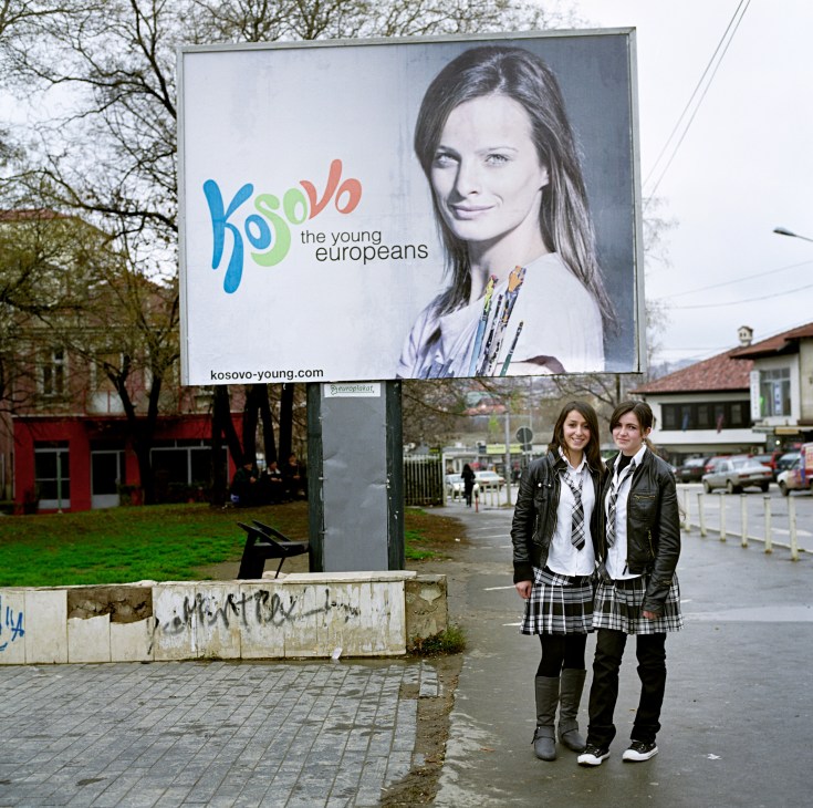 Two teenage girls standing beneath a large billboard for Kosovo's 'The Young Europeans' branding campaign.