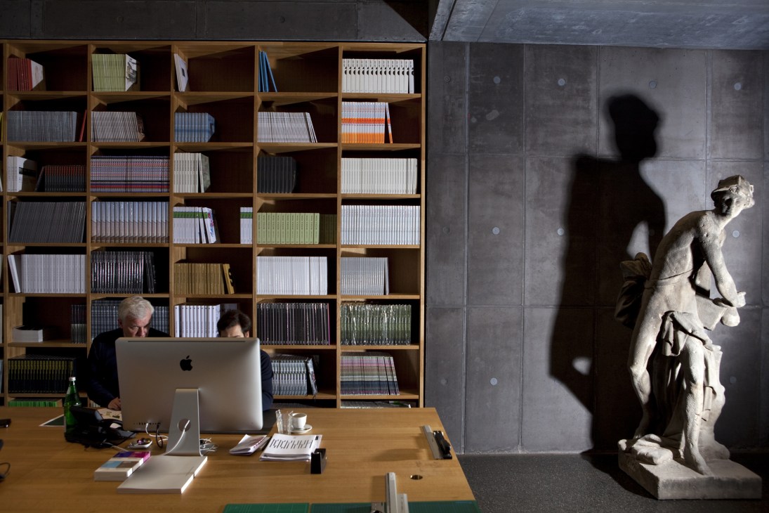 Two people work at a wooden desk in front of a large wooden bookshelf filled with colorful book spines, with a classical stone statue casting a shadow on the gray wall beside them.