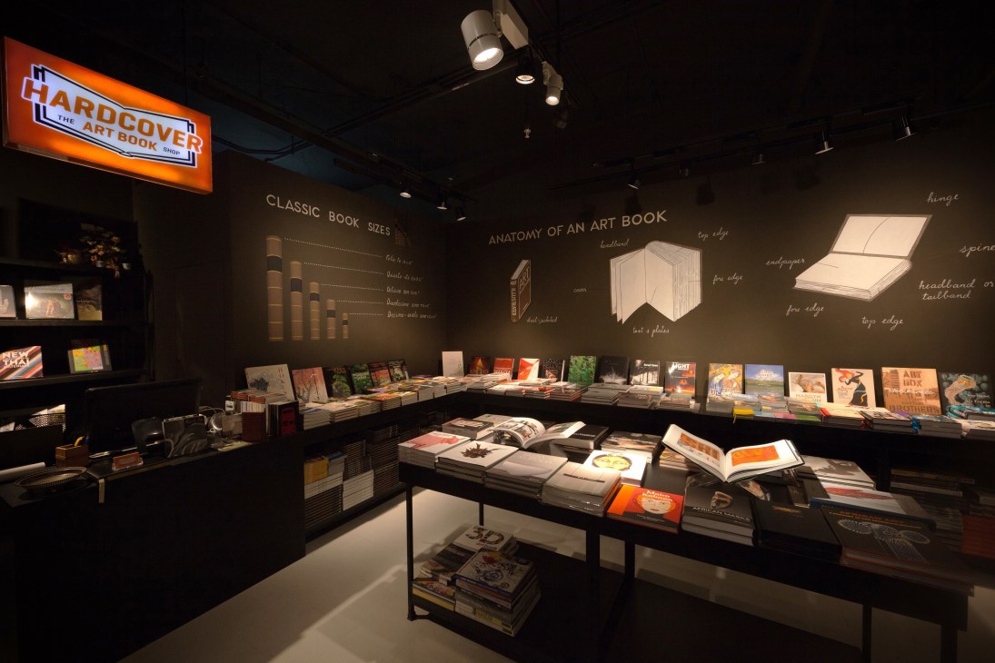 Hardcover bookshop interior in Bangkok with illuminated orange signage, black walls, and displayed art books on tables and shelves.