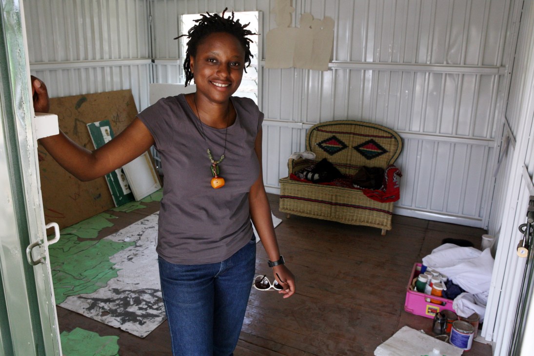 Kenyan artist Ato Abe smiles while holding artwork in her studio with African masks and materials visible.