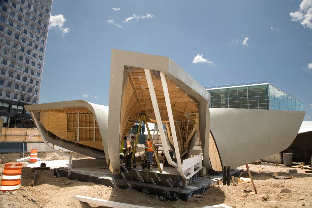 The New Amsterdam Pavilion under construction in New York with curved white concrete forms and wooden interior framework, workers visible inside.