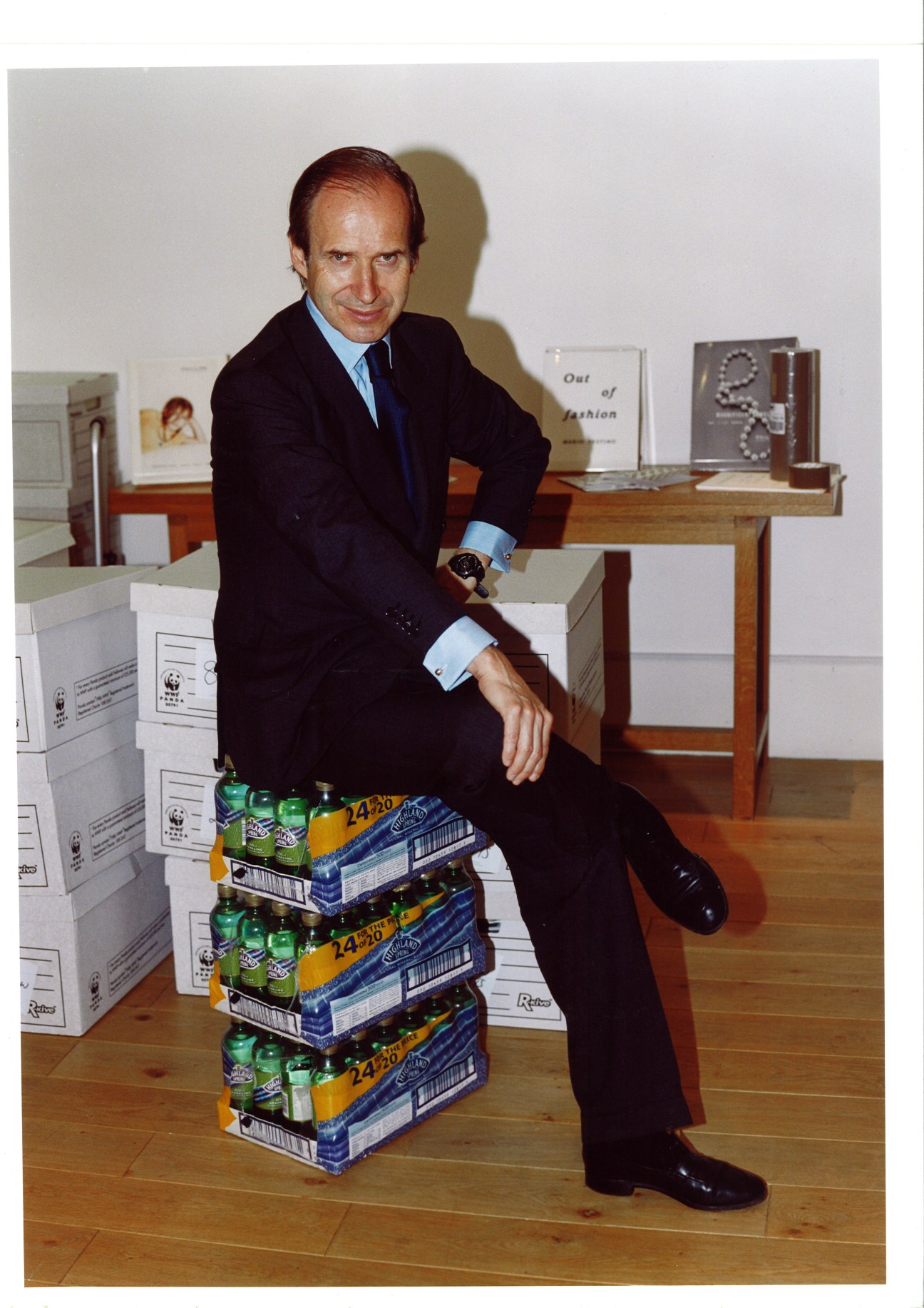 Man in dark suit and light blue shirt sitting on stacked beer cases in an art gallery.