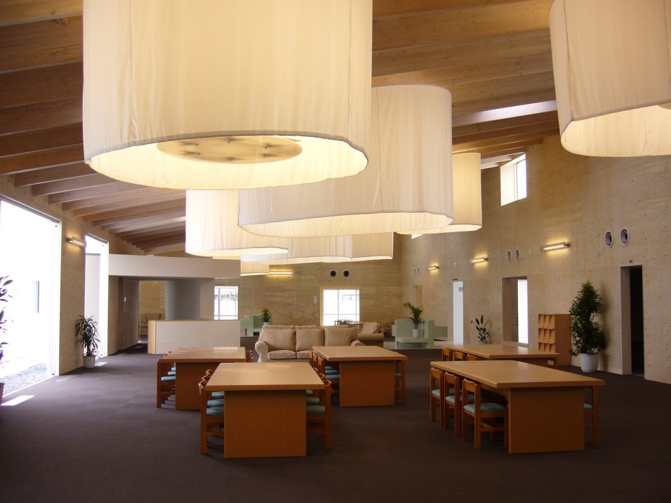 Interior of an international school with wooden ceiling beams, cylindrical pendant lights, wooden tables and chairs, and natural daylight from windows.