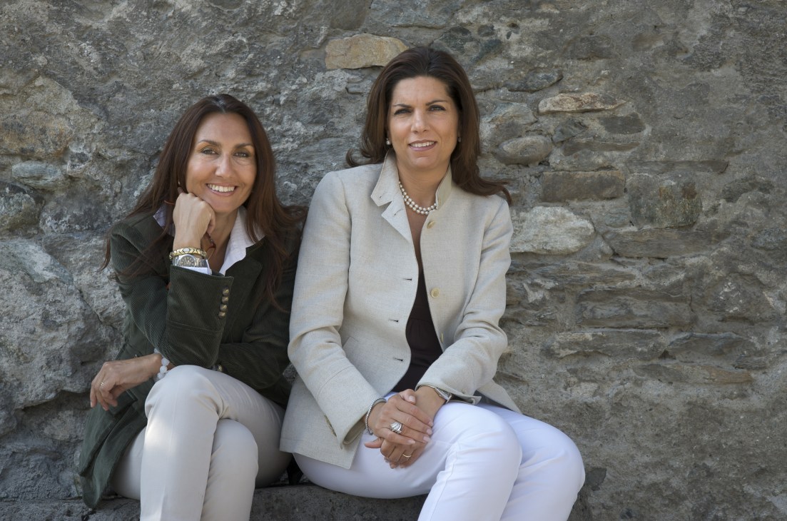 Two women sit against a stone wall, smiling at the camera in professional attire.