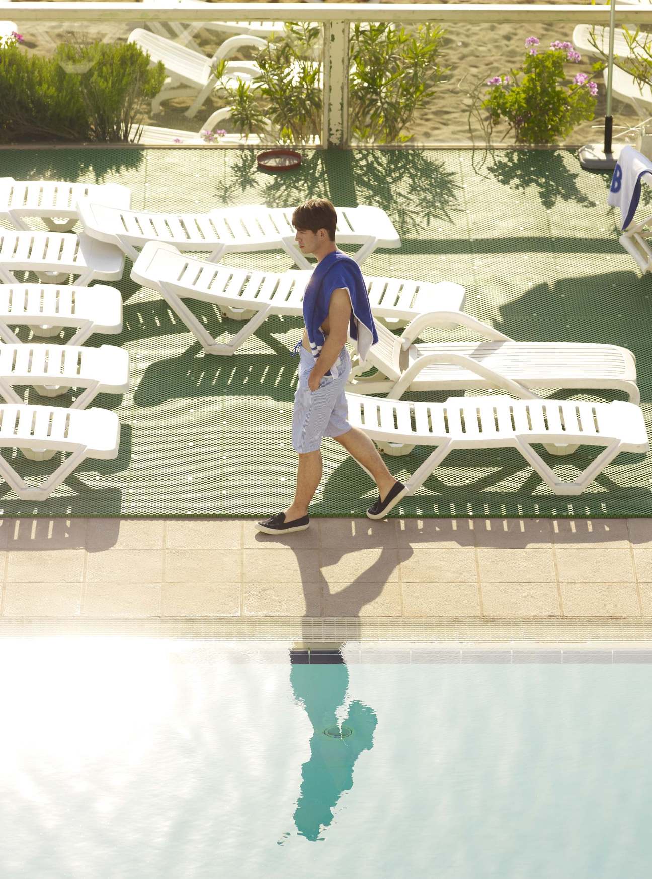 Man wearing blue shirt and light grey swimming shorts walks poolside at Club Natació Barcelona.