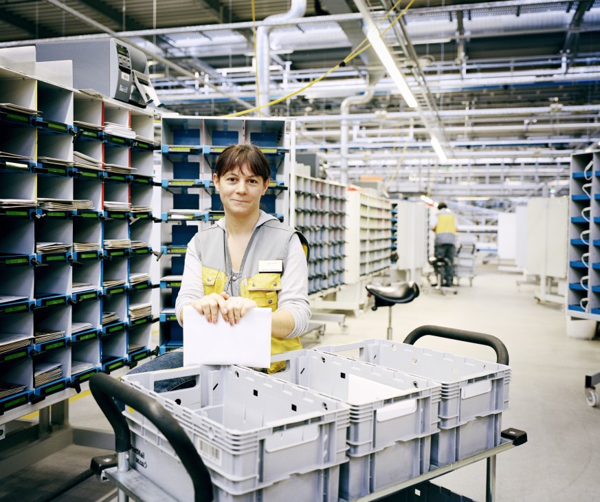 Worker in yellow vest sorting mail at automated sorting facility with high bay racking system.