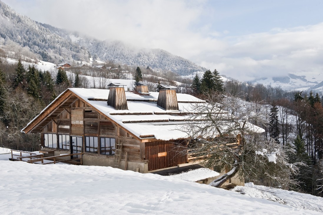 A renovated Alpine barn with wooden and stone exterior sits snow-covered on a mountainside, surrounded by snow-laden evergreens and distant peaks.