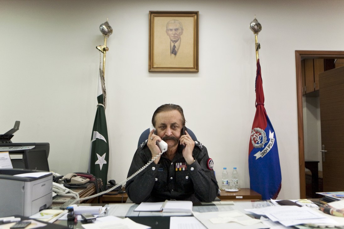 Waseem Ahmed, Karachi's police chief, speaks on two phones simultaneously at his desk in his office, flanked by Pakistani and police flags.