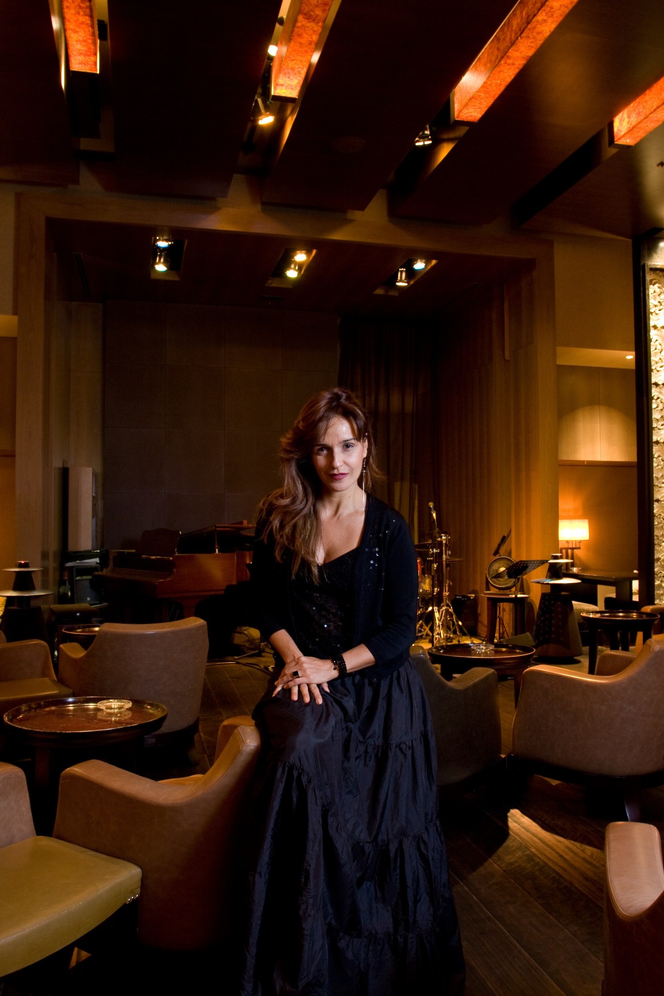Brazilian singer Vanessa Falabella stands in an elegant hotel lounge with a grand piano and jazz club ambiance.