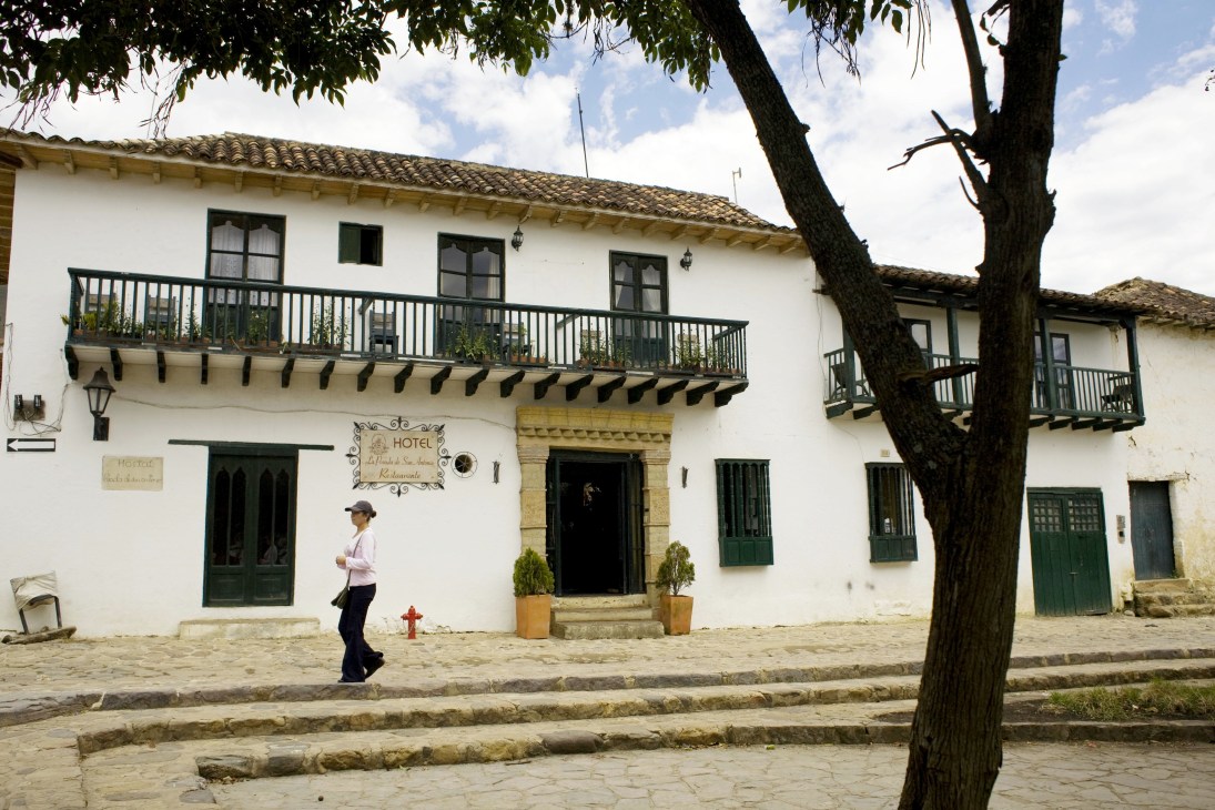 Colonial hotel building with white walls, dark green doors and shutters, wooden balconies, and a large tree in Villa de Leyva town centre.