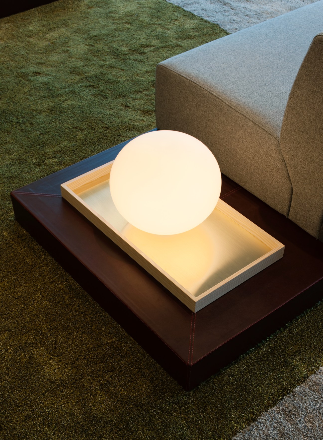 Beige upholstered sofa with integrated glowing spherical lamp on wooden base next to green rug.