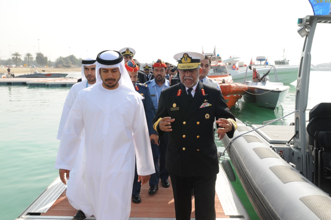 Sheikh Sultan Bin Tahnoon Al Nahyan and Major General Obaid Al Ketbi walk on a dock at IDEX with naval vessels and officials in the background.