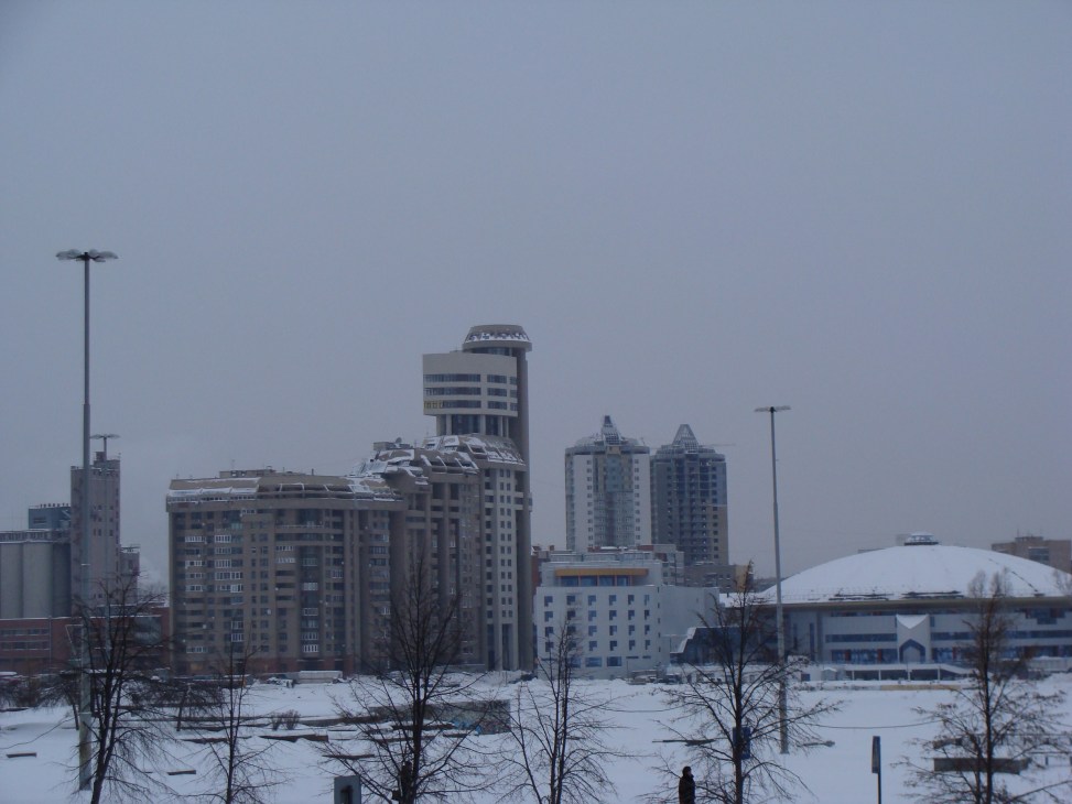 Snow-covered Ekaterinburg cityscape with residential tower blocks and public buildings under overcast sky.