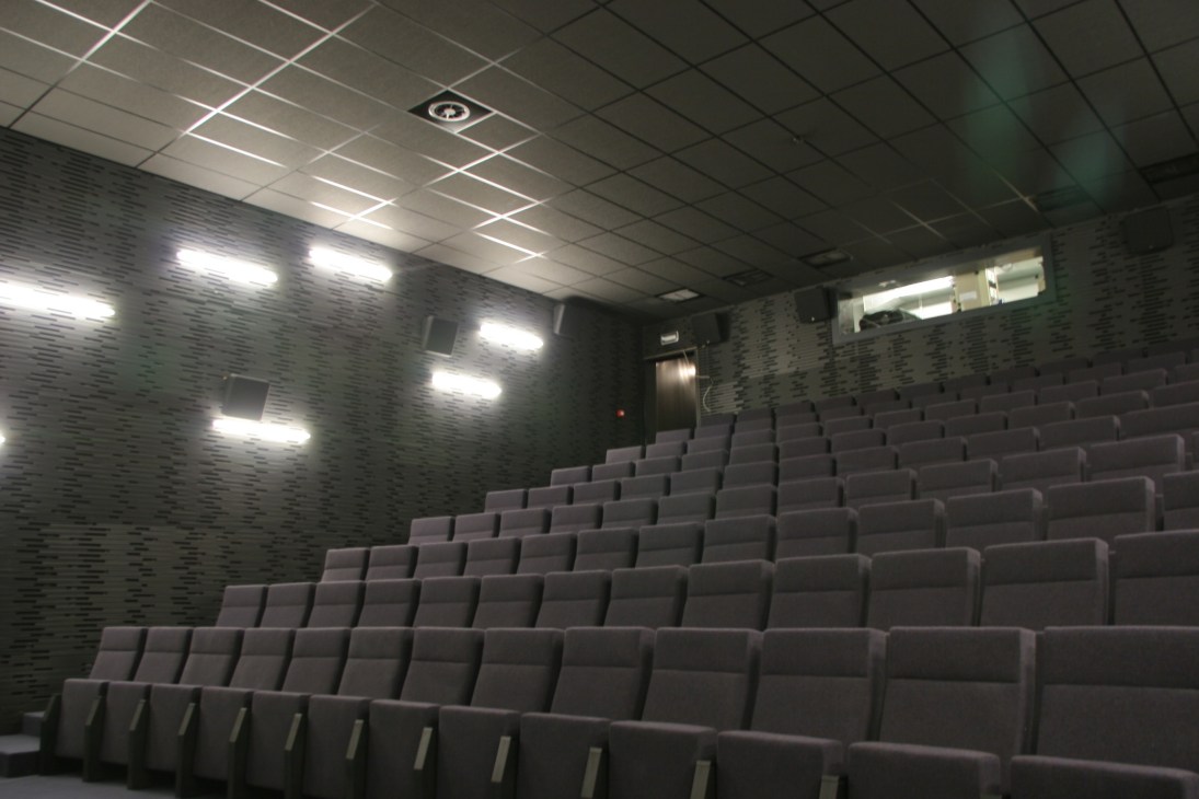 Modern cinema screening room with gray upholstered seats arranged in tiered rows, textured walls, and contemporary lighting fixtures.