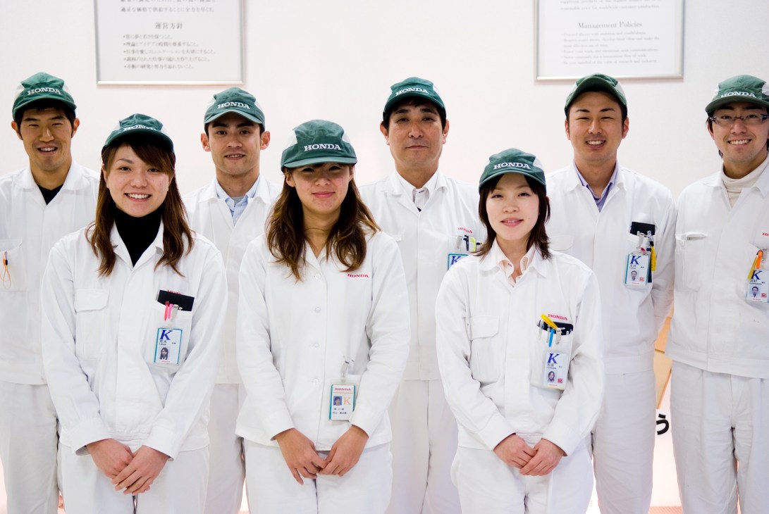 Nine Honda factory workers in white uniforms and green Honda caps posing together indoors.