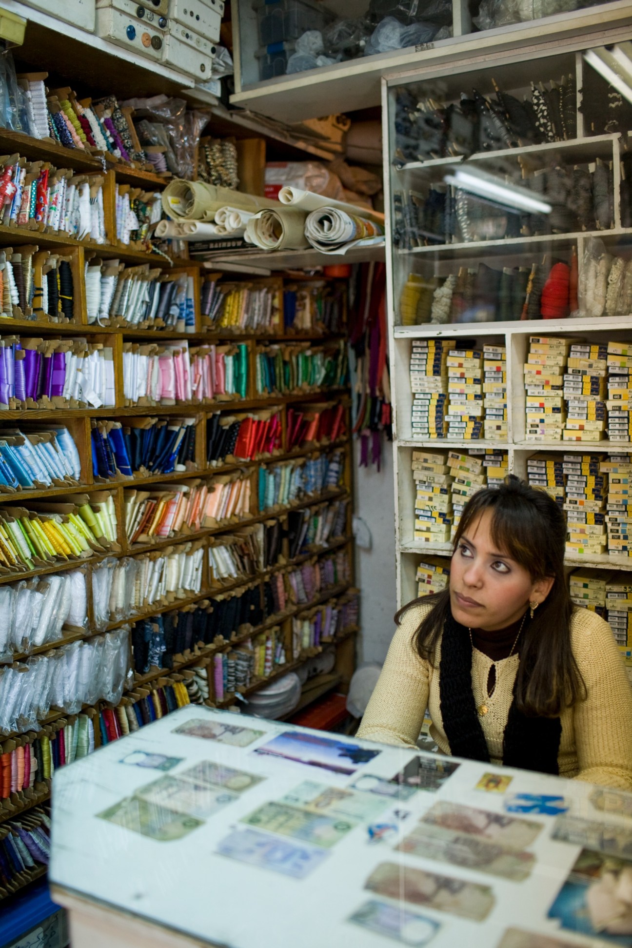 Woman sits at counter in Helio haberdashery surrounded by shelves of colorful ribbons, threads, fabrics and sewing supplies.