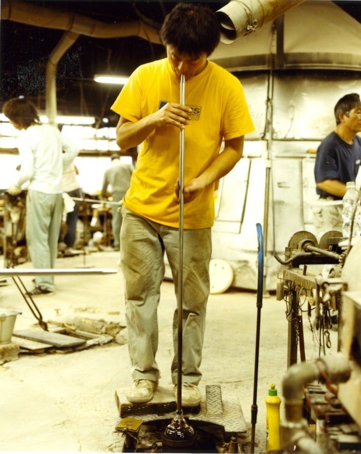 Glass blower in yellow shirt working with molten glass at Shotoku's Tokyo factory.
