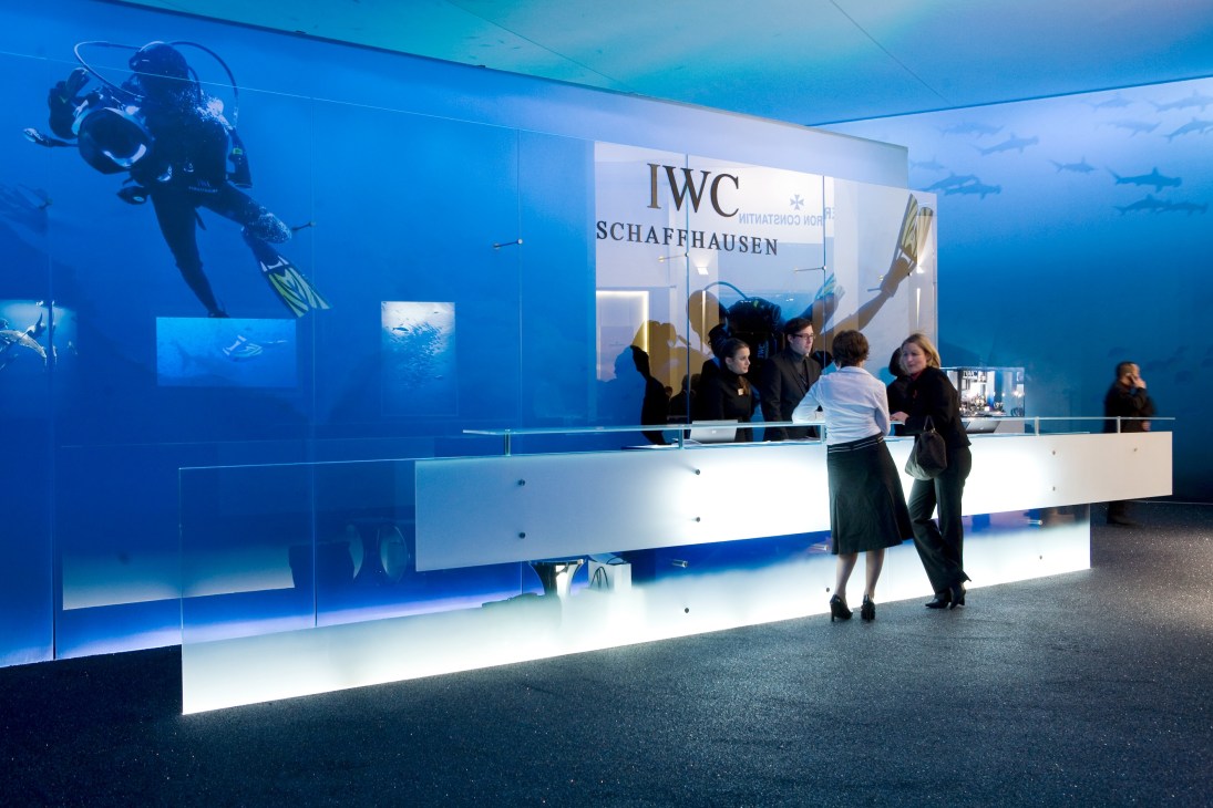 IWC Schaffhausen booth at watch fair featuring underwater diving imagery and blue-lit display with visitors at counter.