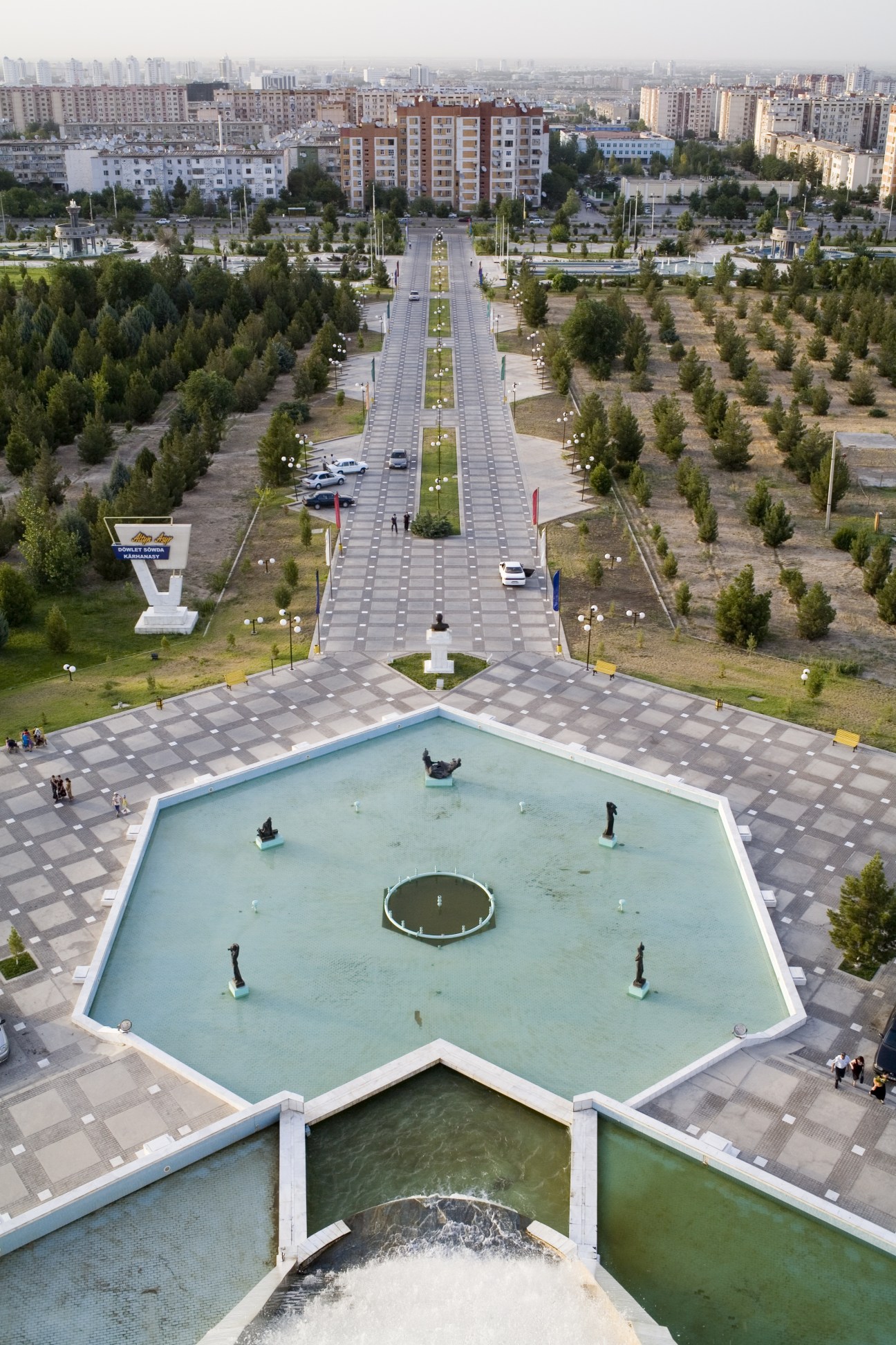 Aerial view of Independence Park in Ashgabat with a large fountain, tree-lined pathway, and Soviet-era apartment buildings in the background.