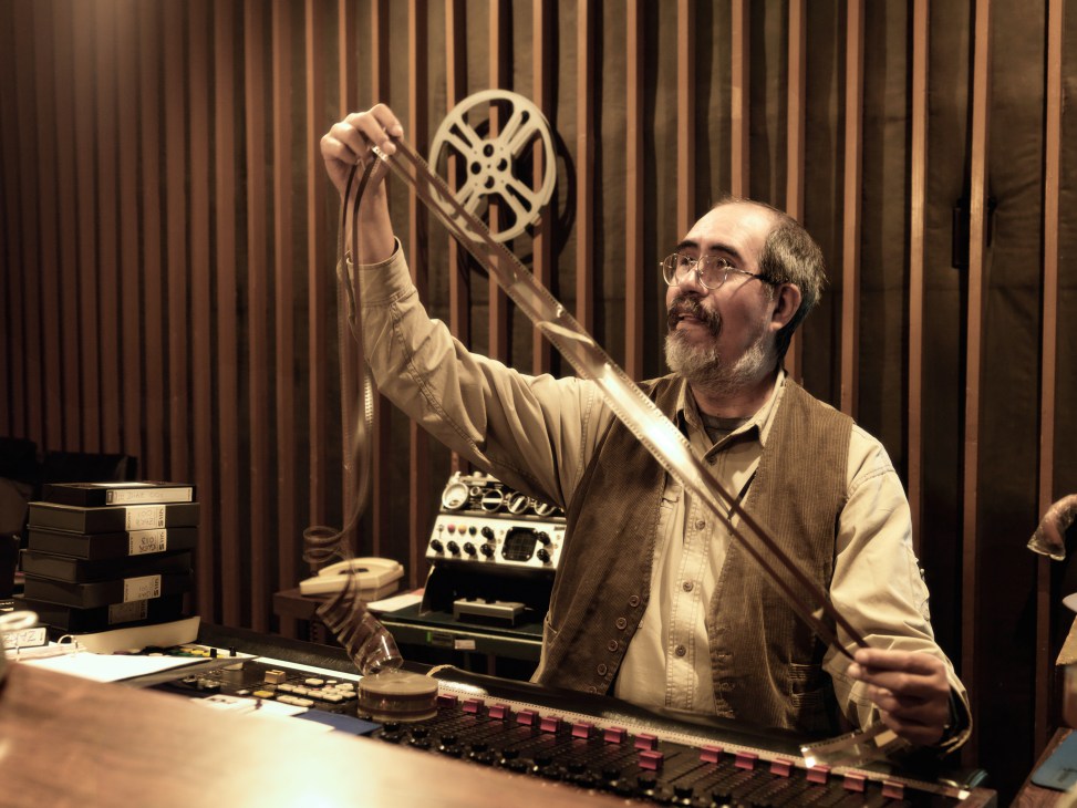 Man holding film reel at Estudios Churubusco sound archives with equipment and vintage curtains behind him.