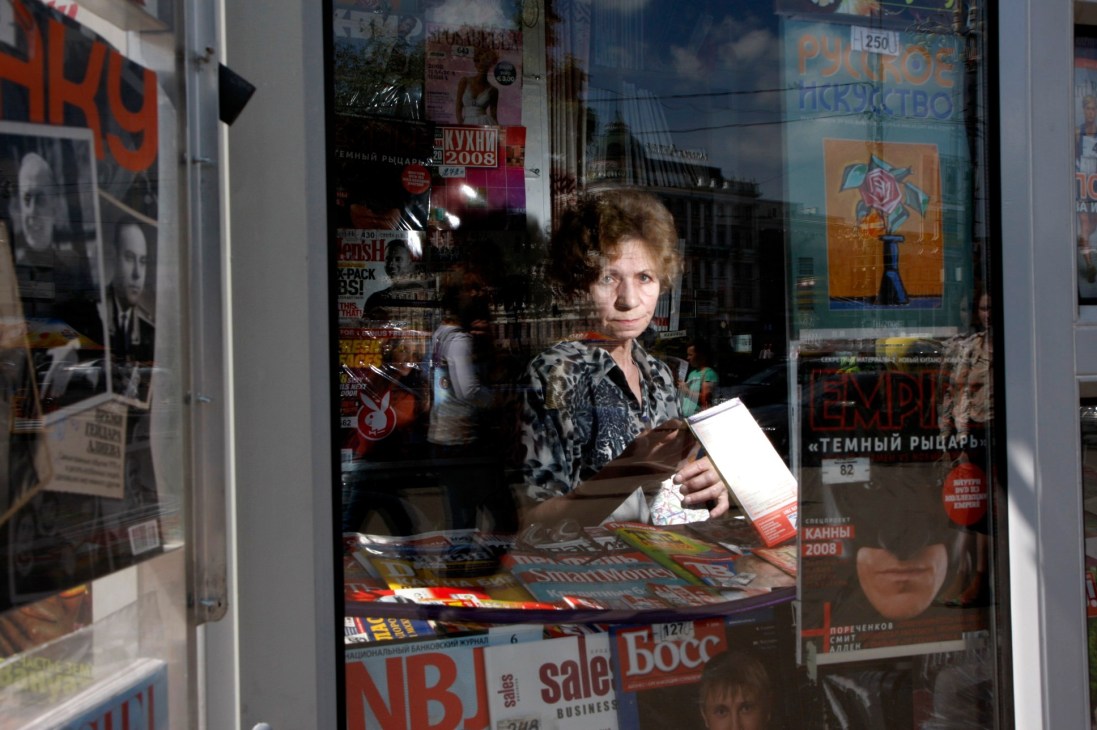 Larisa Petrova stands inside her newspaper and magazine kiosk on Pushkin Square in Moscow, holding a magazine.