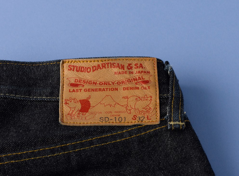 Close-up of a Studio D'Artisan denim jeans label showing red text on tan leather against dark indigo fabric.