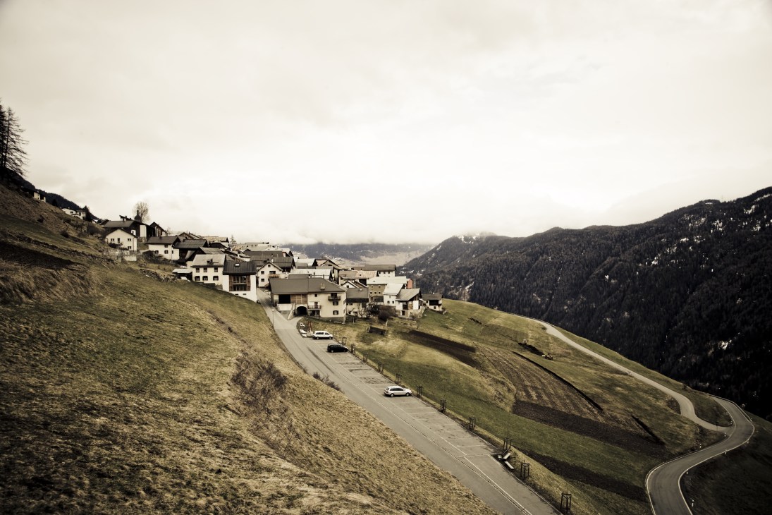 Alpine village of Tschlin nestled in the Engadine valley with traditional Swiss buildings on a hillside surrounded by forested mountains.