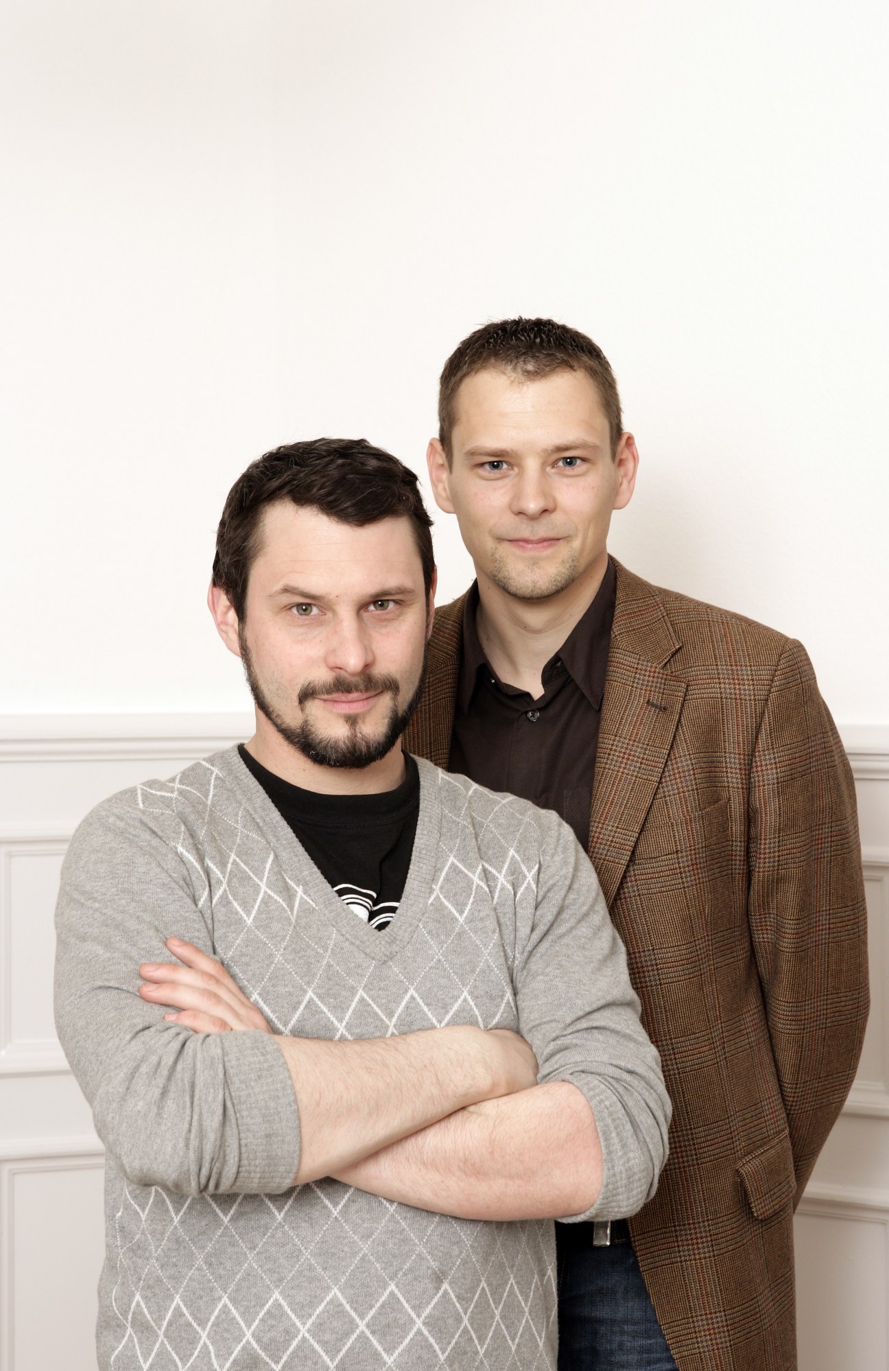 Two men pose for a portrait against a white background; the man on the left wears a grey patterned sweater over a black shirt, while the man on the right wears a brown checked blazer.