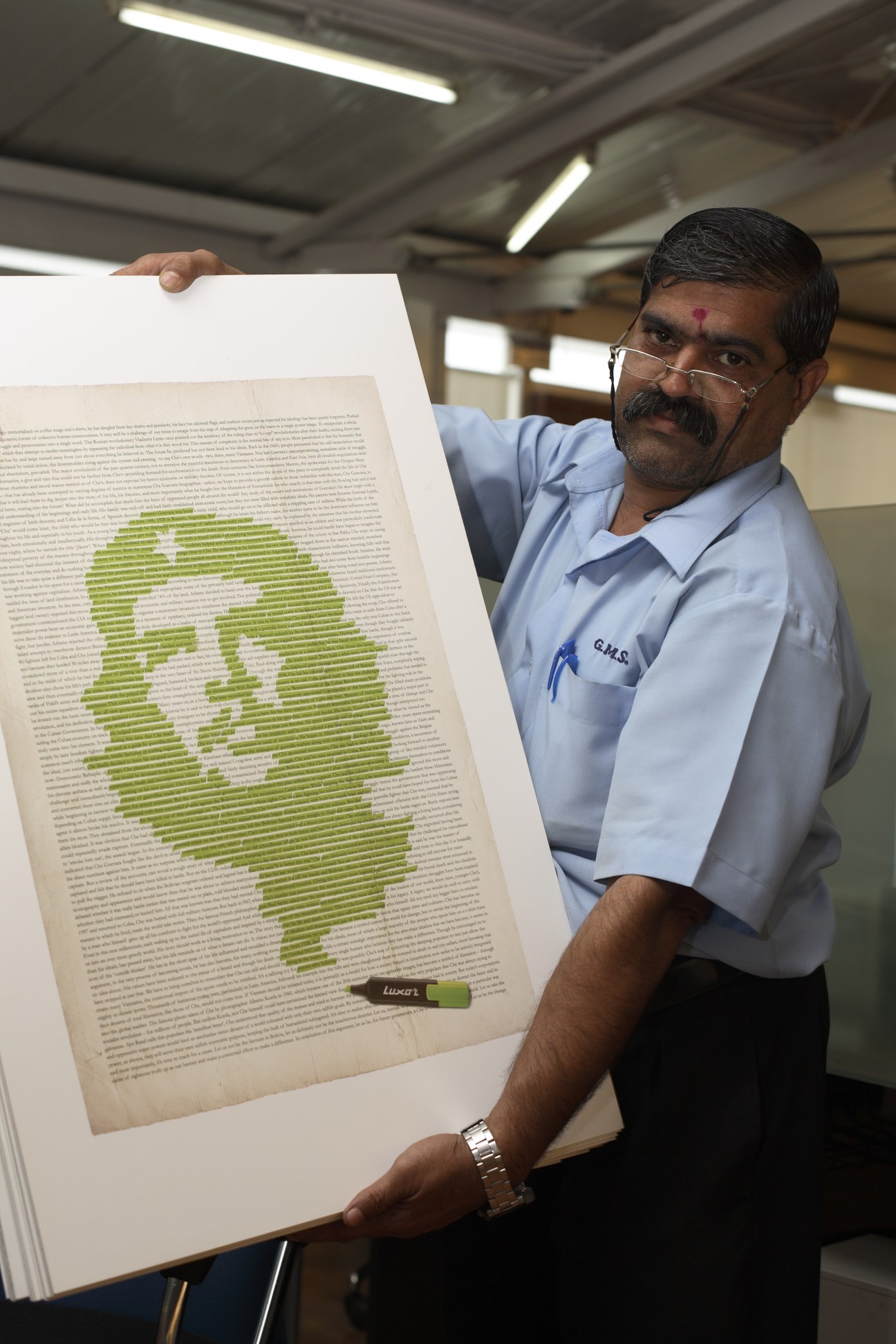 Man holding print advertisement with Che Guevara portrait highlighted in green Luxor ink over newsprint text.