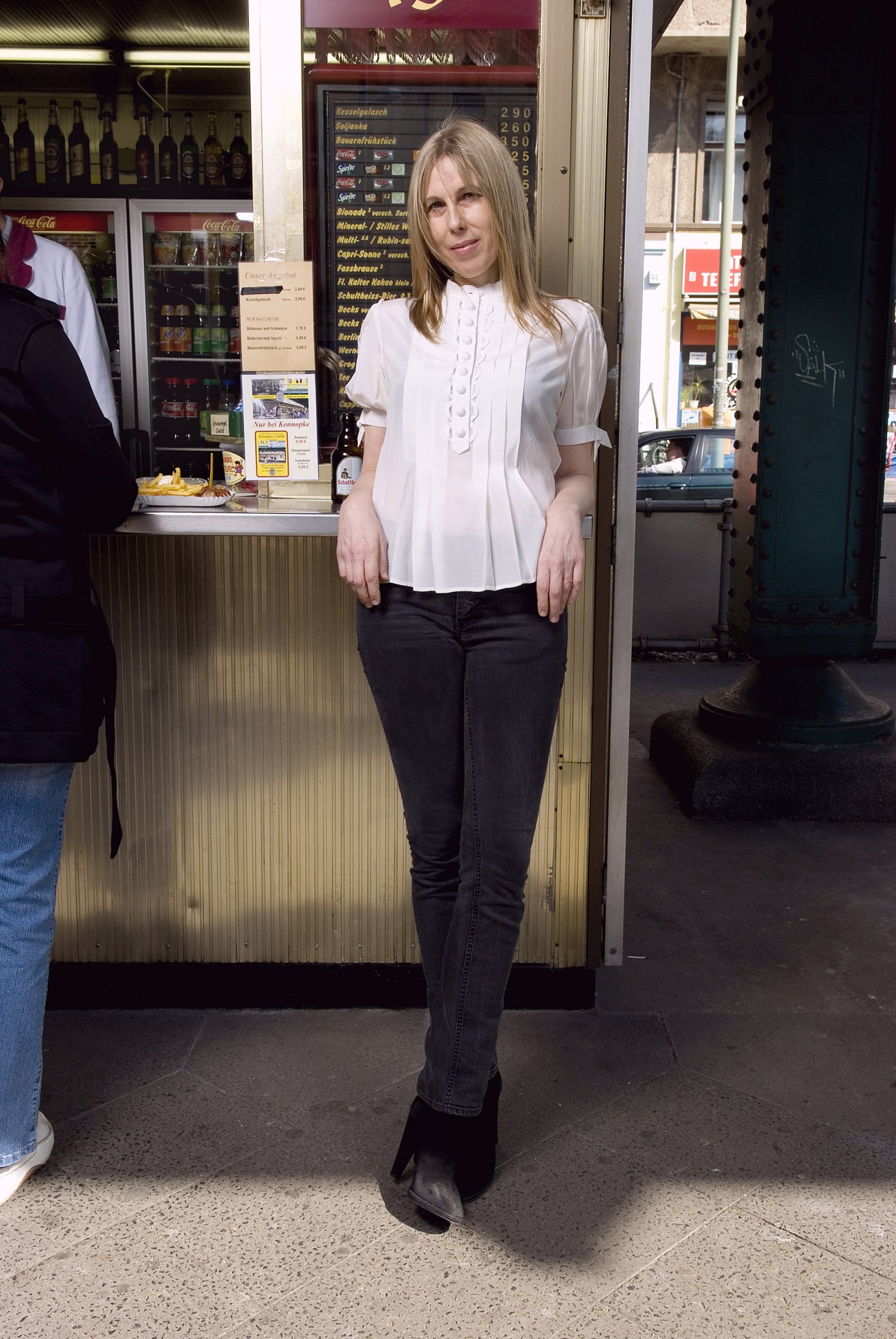 Angelika Taschen stands outside Konnopke's currywurst stand in Berlin, wearing a white blouse and dark jeans.