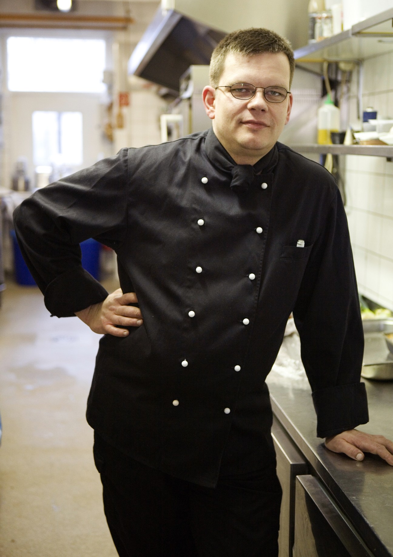 Chef Christian Drewetsch standing in a kitchen wearing a black double-breasted chef's jacket with white buttons.