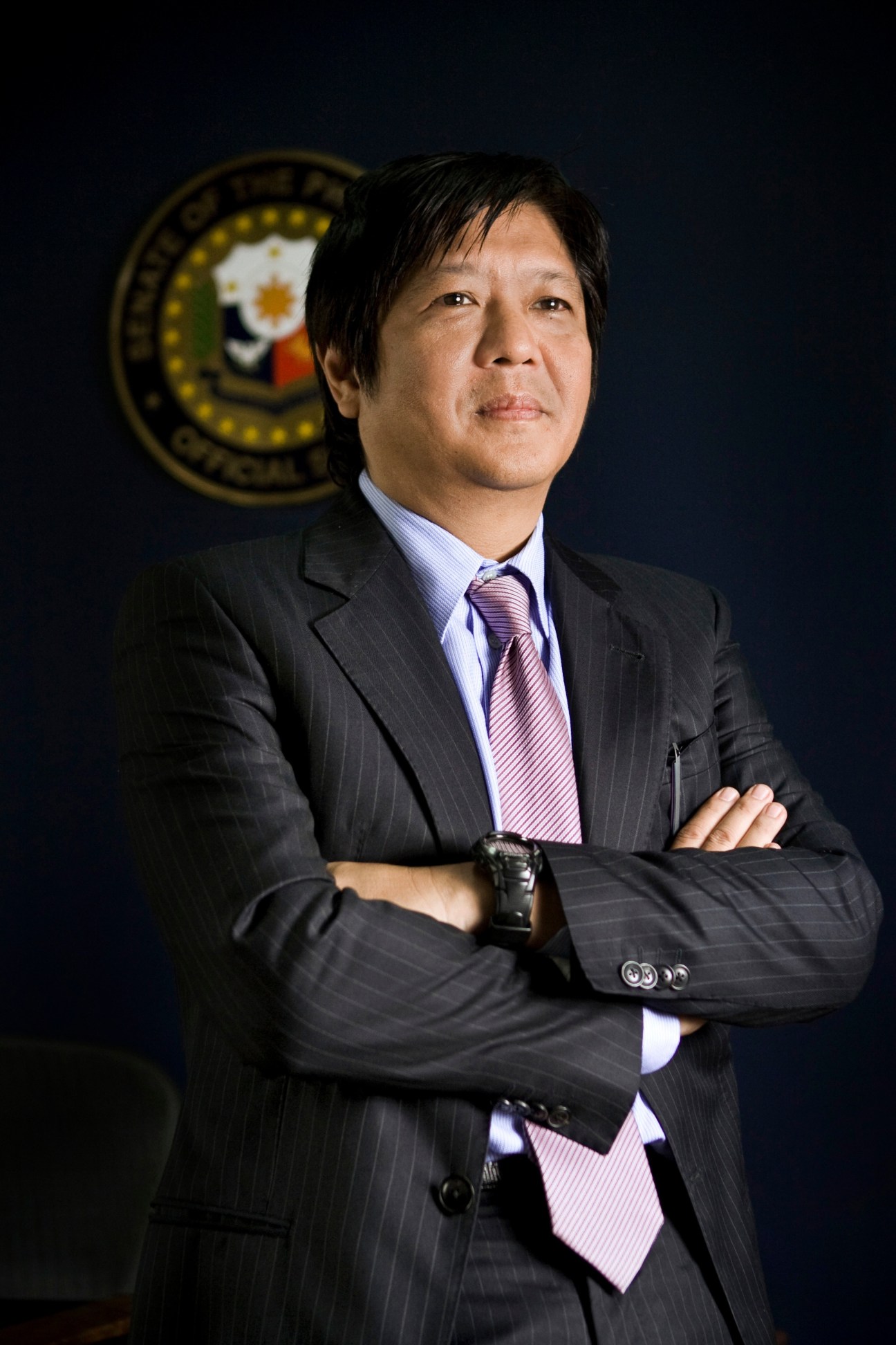 Ferdinand Marcos Jr. in dark pinstriped suit and purple striped tie poses with arms crossed before Philippine seal.