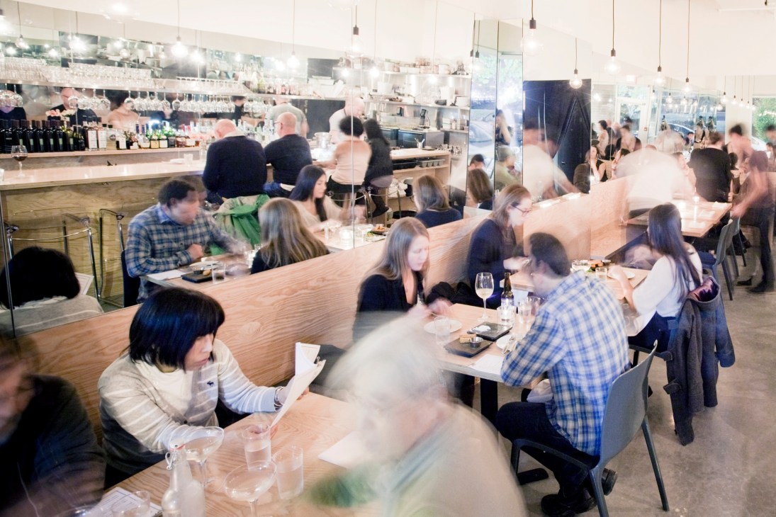 Interior of The Parker restaurant in Vancouver with diners seated at wooden tables and an open kitchen.