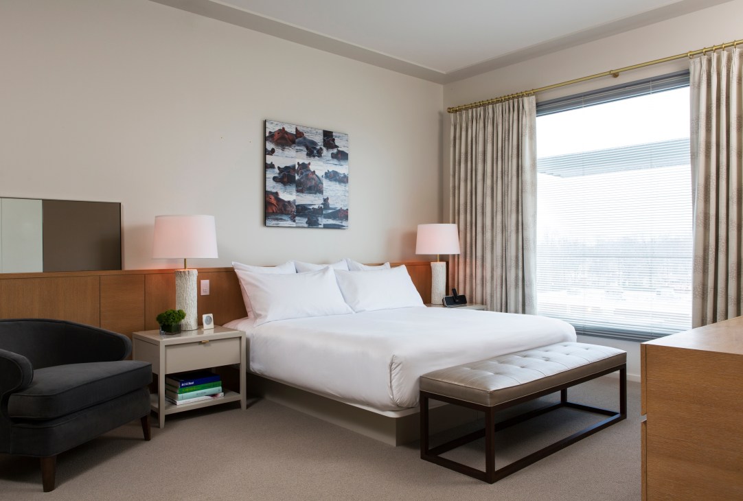 Modern hotel bedroom with white bed, wood furniture, pink lamps, and large windows with curtains.