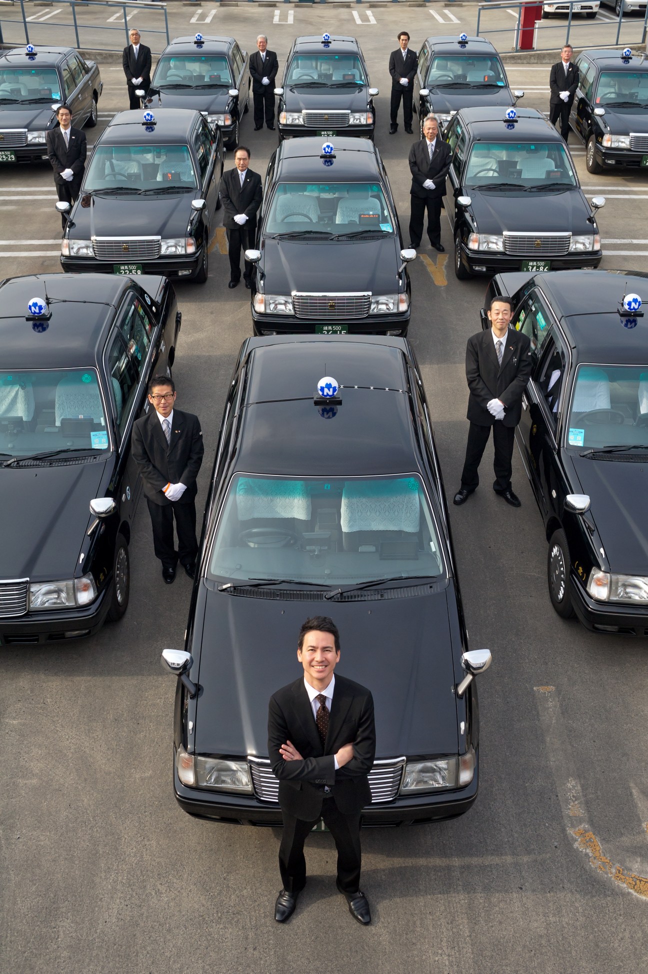 Ichiro Kawanabe stands before rows of Nihon Kotsu taxi drivers in black suits with white gloves, posed with their vehicles.