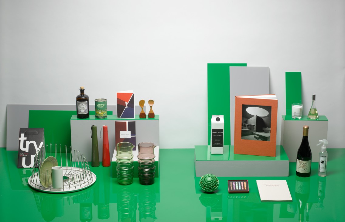 Styled product display featuring gin, wine, umbrellas, dish drainer, books, candles and other curated gift items on green and grey platforms.